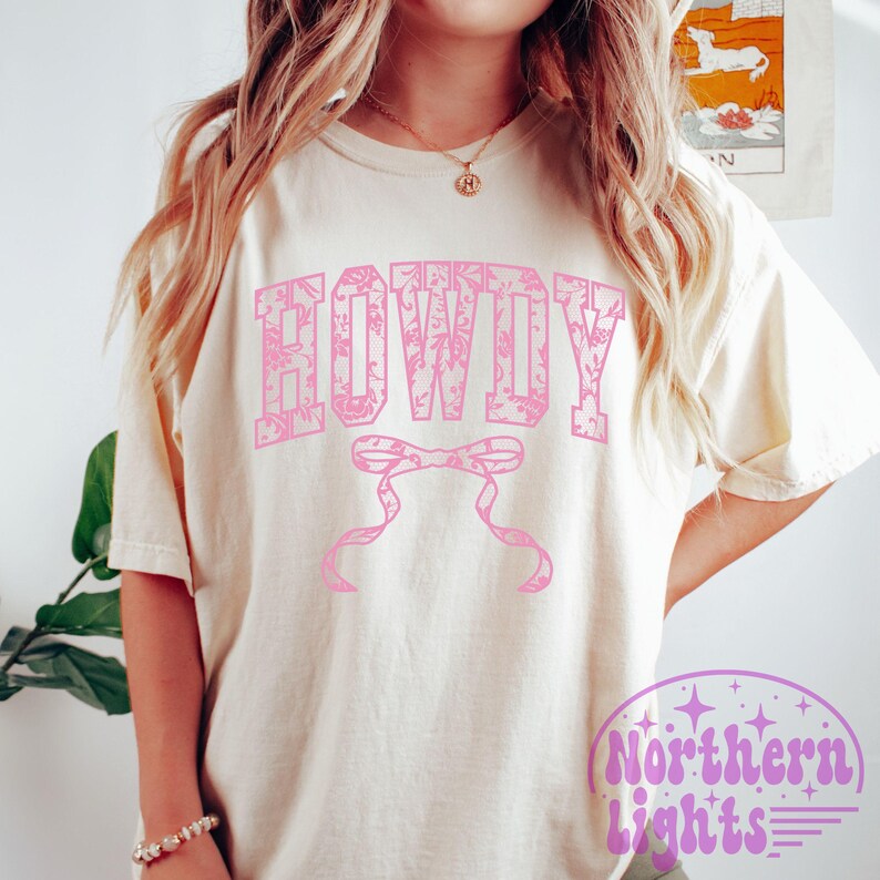 Puede incluir: Una camiseta blanca con un dise&ntilde;o floral rosa que dice "HOWDY" en una fuente floral rosa. La camiseta tiene un lazo rosa con un dise&ntilde;o floral.