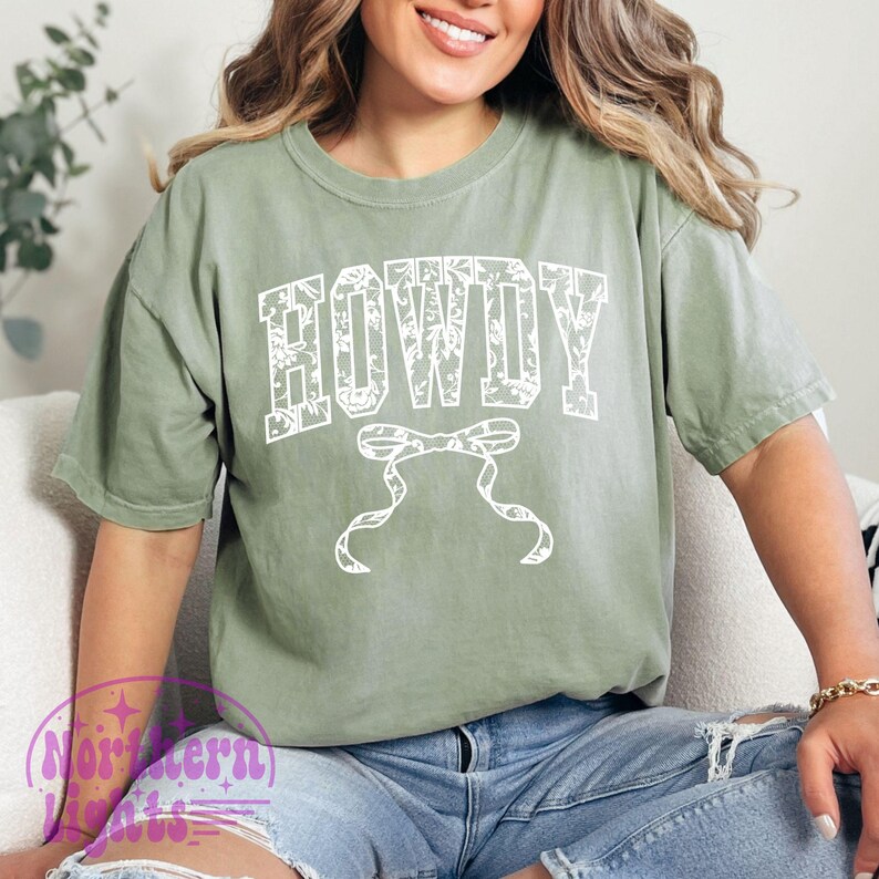 Puede incluir: Una camiseta verde con un estampado floral blanco y la palabra "HOWDY" en blanco con un lazo blanco debajo.