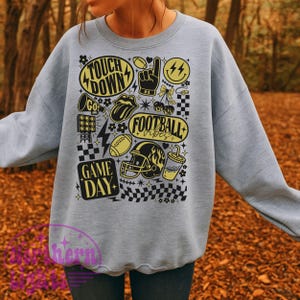May include: A light gray sweatshirt featuring a collection of yellow and black football-themed graphics. The design includes the words "Touch Down", "Football Vibes", and "Game Day", along with footballs, a helmet, and a smiley face.