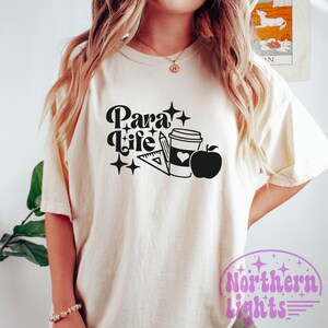 Op de afbeelding: Wit T-shirt met zwarte tekst die "Para Life" zegt en een afbeelding van een koffiebeker, een liniaal, een potlood en een appel.