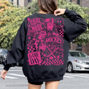 May include: Black sweatshirt with a pink graphic design featuring hockey-themed illustrations and text. The design includes the words "GOAL MODE", "HOCKEY VIBES", and "GAME DAY", along with hockey sticks, a puck, and a helmet.