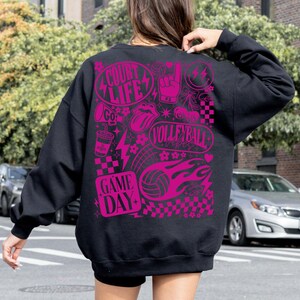 K&ouml;nnte beinhalten: Schwarzes Sweatshirt mit einem leuchtend pinkfarbenen Grafikdesign zum Thema Volleyball. Das Design enth&auml;lt die W&ouml;rter "Court Life", "Volleyball Vibes" und "Game Day". Das Sweatshirt hat einen Rundhalsausschnitt und lange &Auml;rmel.