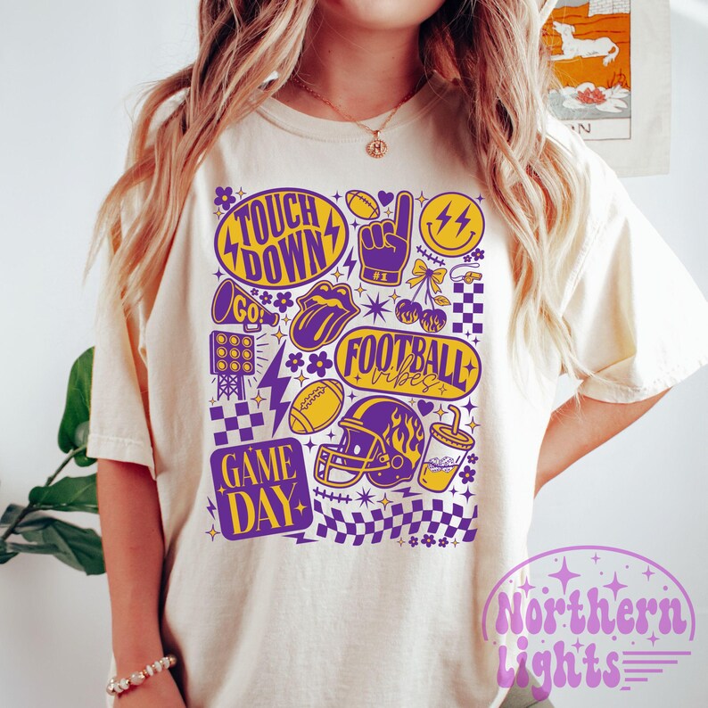 May include: Cream-colored t-shirt with a purple and yellow football-themed graphic. The design includes the words "Touchdown," "Football Vibes," and "Game Day," along with footballs, a helmet, and a lightning bolt.