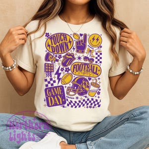 May include: A cream-colored t-shirt featuring a vibrant graphic design with football-themed elements. The design includes the words "Touch Down", "Game Day", and "Football Vibes", along with footballs, a helmet, and a thumbs-up symbol in purple and yellow.