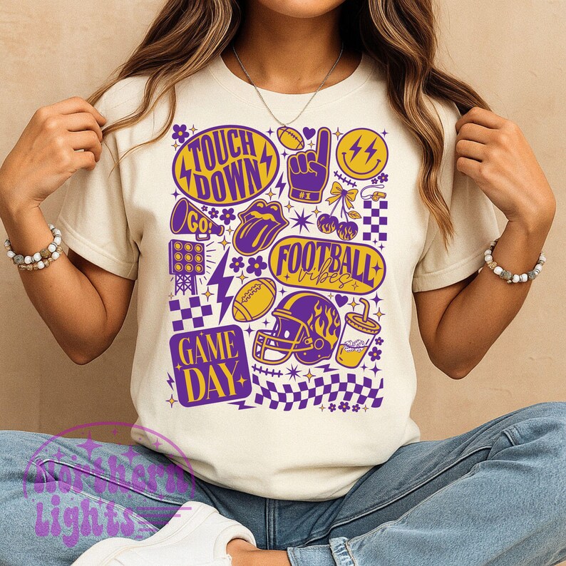 May include: A cream-colored t-shirt featuring a vibrant graphic design with football-themed elements. The design includes the words "Touch Down", "Game Day", and "Football Vibes", along with footballs, a helmet, and a thumbs-up symbol in purple and yellow.
