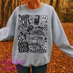 May include: A light gray sweatshirt with a black graphic design featuring basketball-themed elements. The design includes the words "HOOP LIFE," "BASKETBALL VIBES," and "GAME DAY," along with basketballs, a sneaker, and a hand gesture.