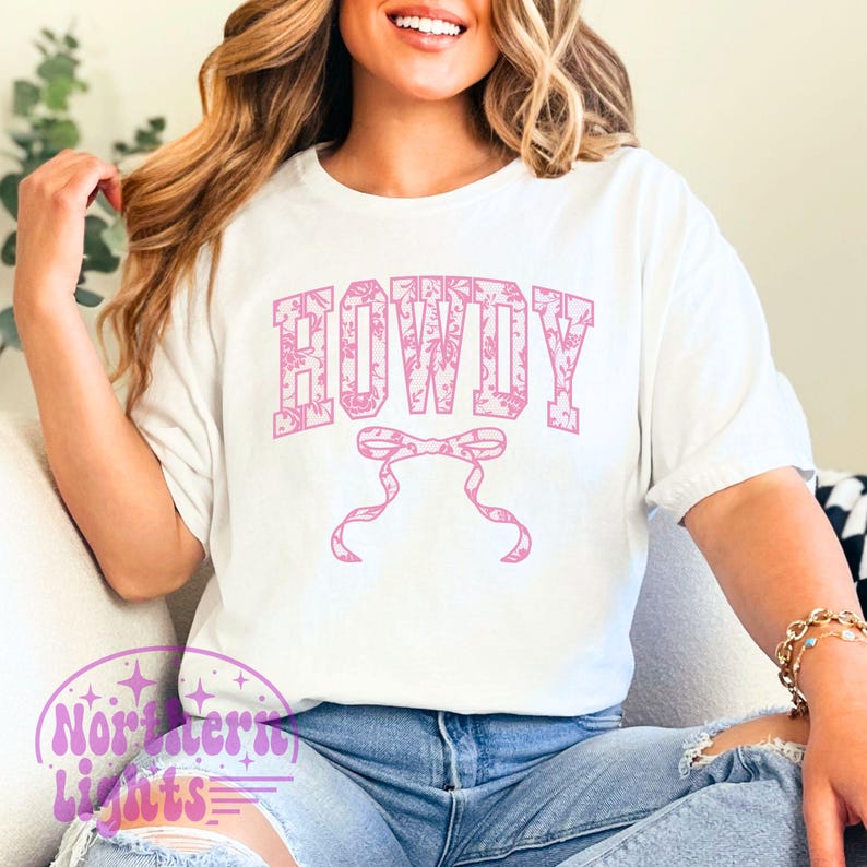 Puede incluir: Camiseta blanca con un dise&ntilde;o floral rosa que dice "HOWDY" en una fuente floral rosa. Un lazo floral rosa est&aacute; debajo del texto.