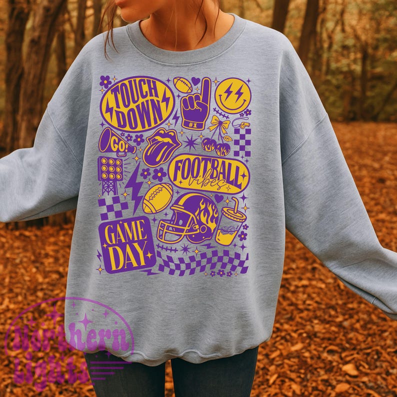 May include: Light blue sweatshirt with a graphic design featuring football-themed elements in purple and yellow. The design includes the words "Touch Down", "Game Day", and "Football Vibes", along with footballs, a helmet, and a thumbs-up.