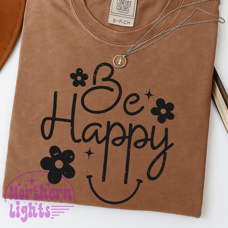 May include: A brown t-shirt with the words "Be Happy" in black script, adorned with flower and star graphics. A silver necklace with a pendant rests on the shirt. The shirt has a crew neck and a small tag at the top.