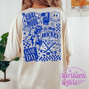 May include: Cream-colored t-shirt with a blue graphic design featuring hockey-themed illustrations and text. The design includes the phrases "GOAL MODE," "HOCKEY vibes," and "GAME DAY," along with hockey sticks, pucks, and other related imagery.