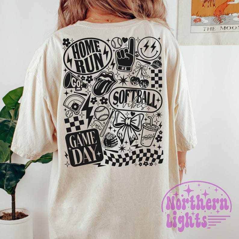 May include: A cream-coloured t-shirt featuring a black and white graphic on the back. The design incorporates the phrases "HOME RUN", "SOFTBALL vibes", and "GAME DAY", alongside baseball-related illustrations. The t-shirt has a loose fit.