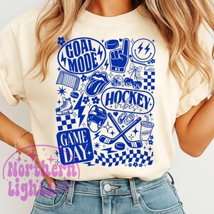 May include: Cream-colored sweatshirt with a blue hockey-themed graphic. The design includes the words "GOAL MODE", "HOCKEY VIBES", and "GAME DAY", along with hockey pucks, sticks, and other related icons. The design is printed in blue.