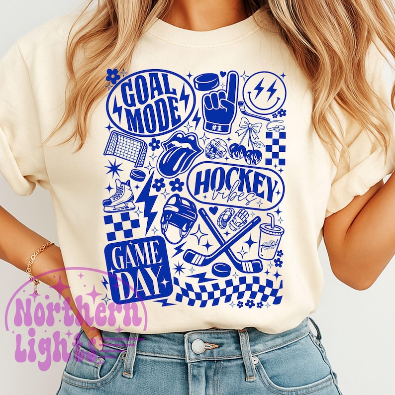 May include: Cream-colored sweatshirt with a blue hockey-themed graphic. The design includes the words "GOAL MODE", "HOCKEY VIBES", and "GAME DAY", along with hockey pucks, sticks, and other related icons. The design is printed in blue.