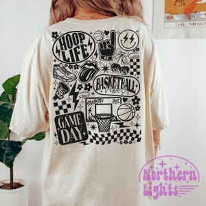 May include: Cream-colored t-shirt with a black basketball-themed graphic. The design includes the words "HOOP LIFE", "BASKETBALL VIBES", and "GAME DAY", along with basketball-related illustrations such as a basketball, hoop, and sneakers.