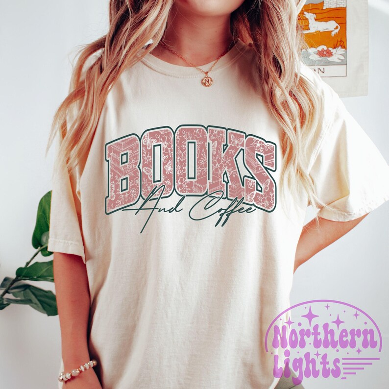 K&ouml;nnte beinhalten: Cremefarbenes T-Shirt mit dem Text "BOOKS And Coffee" in einem rosa Blumendesign. Der Text ist auf der Vorderseite des Shirts gew&ouml;lbt. Die Worte "And Coffee" sind in einer Schreibschrift. Das Shirt hat kurze &Auml;rmel.