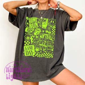 May include: Dark grey t-shirt featuring a neon green softball design. The graphic includes the words "Home Run", "Softball Vibes", and "Game Day", alongside baseballs, lightning bolts, and a bow.