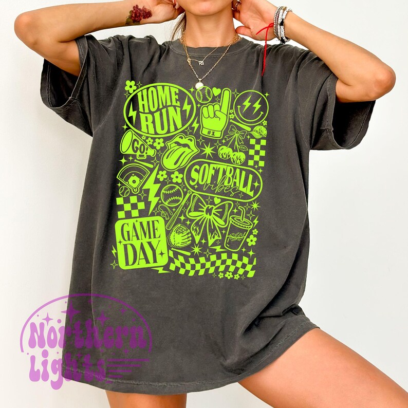 May include: Dark grey t-shirt featuring a neon green softball design. The graphic includes the words "Home Run", "Softball Vibes", and "Game Day", alongside baseballs, lightning bolts, and a bow.
