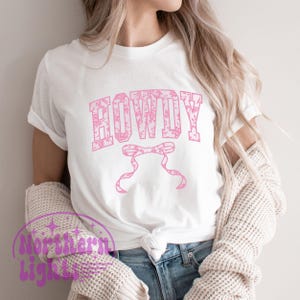 Puede incluir: Una camiseta blanca con un dise&ntilde;o floral rosa que dice "HOWDY" en una fuente floral rosa. La camiseta tiene un dise&ntilde;o de corbata de lazo debajo del texto.