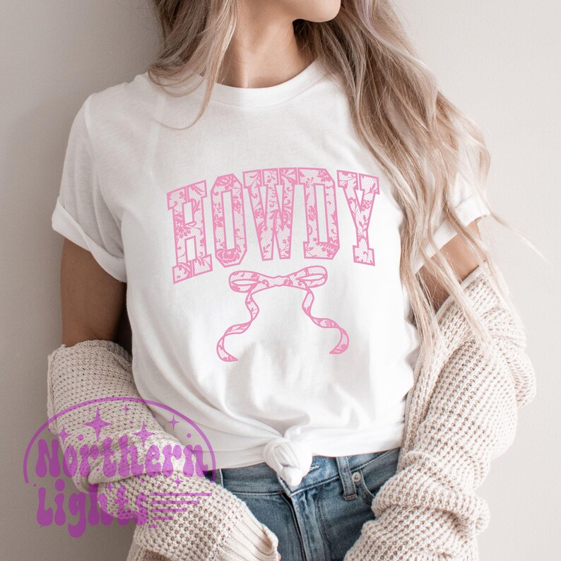 Puede incluir: Una camiseta blanca con un dise&ntilde;o floral rosa que dice "HOWDY" en una fuente floral rosa. La camiseta tiene un dise&ntilde;o de corbata de lazo debajo del texto.