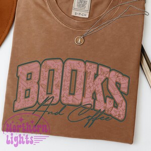 K&ouml;nnte beinhalten: Ein braunes T-Shirt mit dem Schriftzug "BOOKS And Coffee" in einem rosa Blumendesign. Eine silberne Halskette mit Anh&auml;nger liegt auf dem Shirt. Das Shirt hat ein Etikett mit der Aufschrift "CONFORT COLORS".