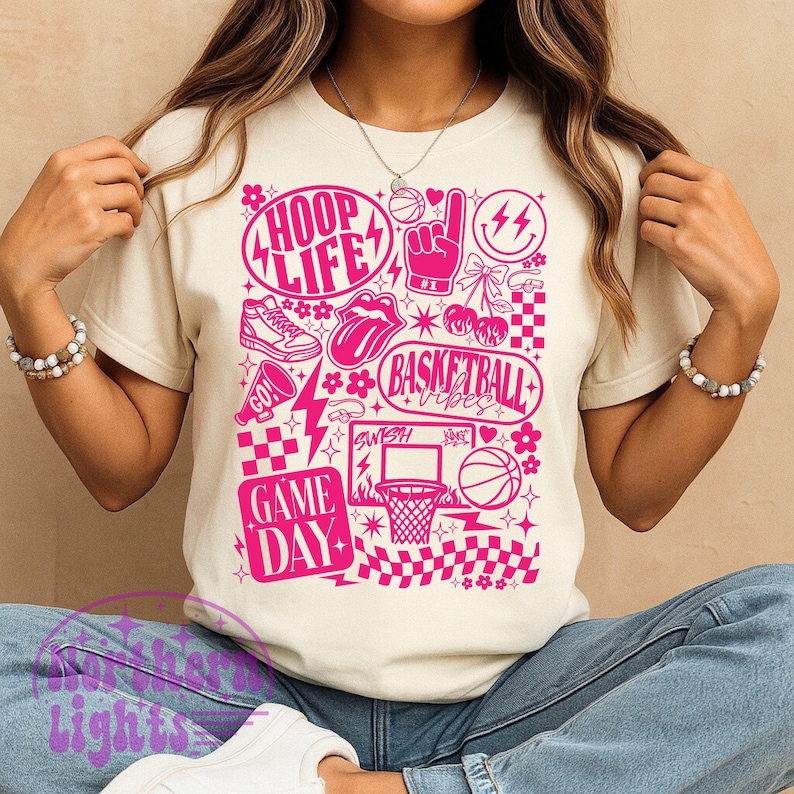 May include: Cream-colored t-shirt with a vibrant pink basketball-themed graphic. The design includes the words "HOOP LIFE," "BASKETBALL VIBES," and "GAME DAY," along with basketball-related imagery such as a basketball, hoop, and sneakers.