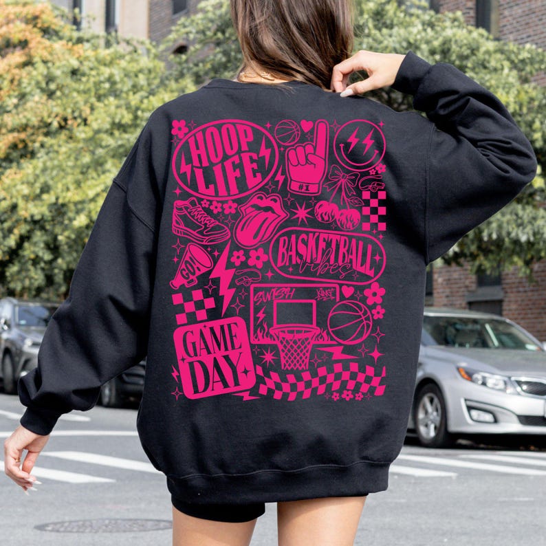 May include: Black sweatshirt with a pink graphic design featuring basketball-themed illustrations and text. The design includes the words "HOOP LIFE," "BASKETBALL VIBES," and "GAME DAY," along with basketballs, a tongue, and a hand gesture.