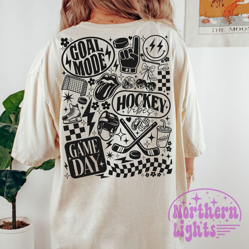 May include: Cream-colored t-shirt with a black and white hockey-themed graphic. The design includes the phrases "Goal Mode," "Hockey Vibes," and "Game Day," along with hockey-related icons such as a puck, skates, and crossed hockey sticks.