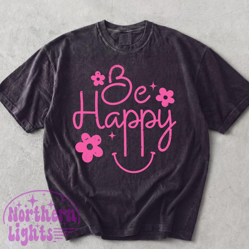 May include: Dark gray t-shirt with the words "Be Happy" in pink script, adorned with flower and star graphics, and a smiling face. The shirt has a vintage, washed appearance.
