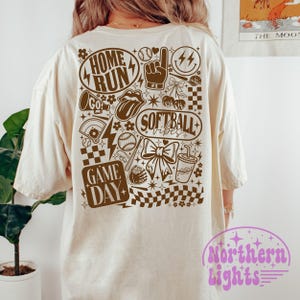 May include: A cream-coloured t-shirt with a brown graphic design featuring baseball and softball elements. The design includes the words "HOME RUN", "GAME DAY", and "SOFTBALL vibes". Other graphics include a baseball, a bow, and a lightning bolt.