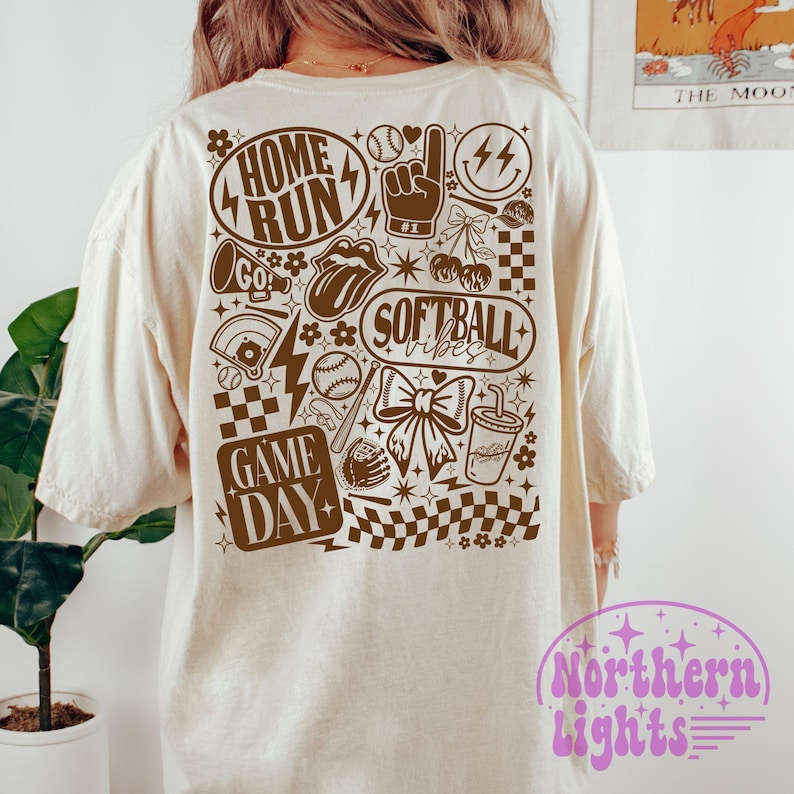 May include: A cream-coloured t-shirt with a brown graphic design featuring baseball and softball elements. The design includes the words "HOME RUN", "GAME DAY", and "SOFTBALL vibes". Other graphics include a baseball, a bow, and a lightning bolt.