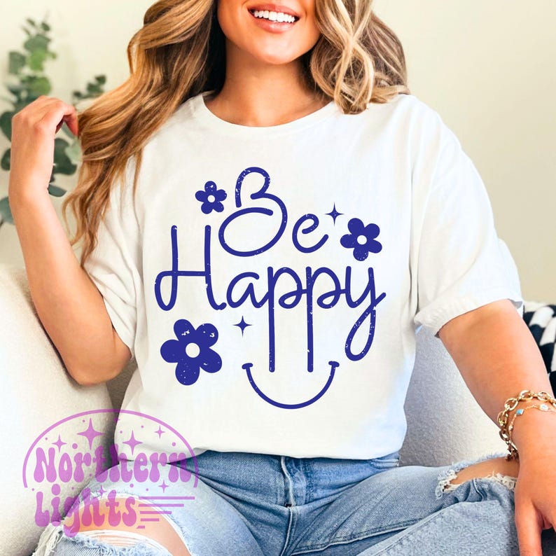May include: White t-shirt with the phrase "Be Happy" in dark blue script, adorned with flower and star graphics, and a smiling face. The shirt is a casual, everyday wear item.