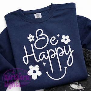 May include: Navy blue sweatshirt with the words "Be Happy" in white script, accompanied by white flower and star graphics. The design includes a smiling face. The sweatshirt is folded.