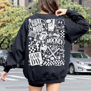 May include: Black sweatshirt with white graphic designs related to hockey. The design includes the words "GOAL MODE", "HOCKEY VIBES", and "GAME DAY", along with hockey-related images such as a puck, skates, and a helmet.