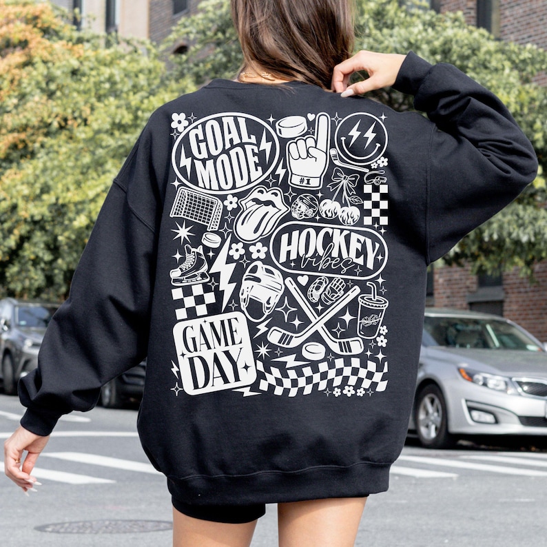 May include: Black sweatshirt with white graphic designs related to hockey. The design includes the words "GOAL MODE", "HOCKEY VIBES", and "GAME DAY", along with hockey-related images such as a puck, skates, and a helmet.