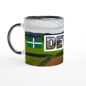 May include: A ceramic mug with a black handle and interior featuring a wrap-around landscape print of green fields, a red soil patch, and a cloudy sky. The design includes a green flag with a white cross and the text DEVO in bold letters.