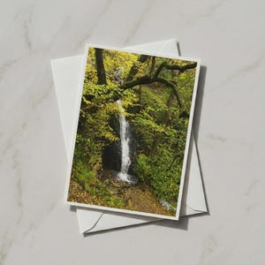 Op de afbeelding: Een rechthoekige wenskaart met een foto van een smalle waterval die langs een donkere, rotsachtige klif naar beneden stroomt, omringd door dicht, levendig groen en geel herfstgebladerte, met een witte envelop direct eronder geplaatst.