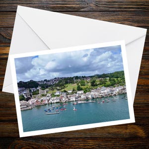 May include: A rectangular greeting card featuring a high-angle photograph of a coastal town with white buildings, a stone church, and numerous small boats floating on blue water under a cloudy sky, accompanied by a white envelope.