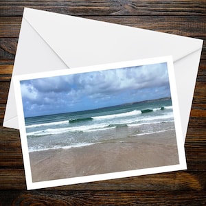 Puede incluir: Una tarjeta de felicitación rectangular que presenta una fotografía de una playa con arena marrón clara, olas de mar turquesa rodando hacia la orilla y un cielo azul lleno de nubes blancas dispersas, presentada con un sobre blanco.