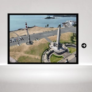 May include: An aerial view of a coastal park featuring a tall stone monument with a globe top, a red and white striped lighthouse, and a parking area with several cars. The landscape includes grassy areas, paved walkways, and a view of the ocean.