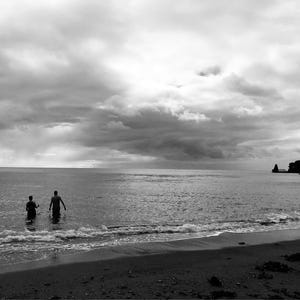 May include: A black and white landscape featuring two figures standing in the ocean water near a sandy beach. The sky is filled with heavy, dark clouds above the horizon, and a dark, rocky cliffside is visible on the right side of the frame.