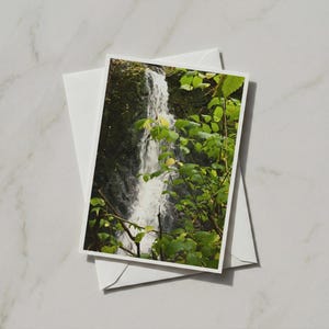 Op de afbeelding: Een rechthoekige wenskaart met een foto van een waterval omringd door weelderig groen gebladerte en donkere rotsformaties, rustend op een effen witte envelop, die een natuurlijk landschap in verticale oriëntatie vastlegt.