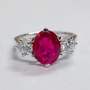 May include: A silver ring with a large oval-shaped red gemstone surrounded by two smaller clear gemstones.