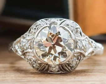 May include: A vintage-style engagement ring with a large, round, clear diamond surrounded by smaller diamonds. The ring is set in platinum and has a delicate, intricate design.