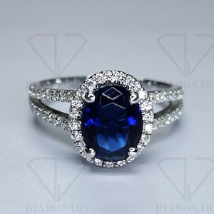 May include: A silver ring with a large oval blue sapphire surrounded by a halo of smaller diamonds. The ring has a split shank with a row of diamonds on each side.