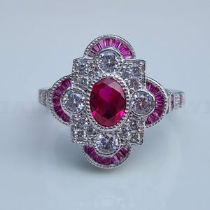 May include: A silver ring with a central oval ruby surrounded by white and pink gemstones. The ring has a geometric design with a cluster of white stones and pink baguette-cut stones. The ring is set against a white background.