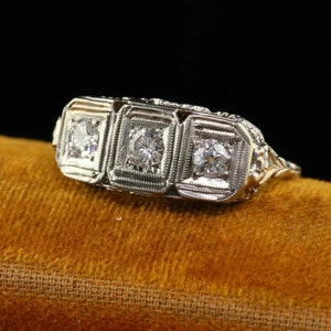 May include: A vintage three-stone diamond ring with a delicate filigree design. The ring is set in platinum and features three round brilliant diamonds.