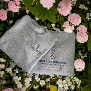 May include: Two gray sweatshirts with the text "Furbies Somerset Pawsitively the Best" and a paw print logo. The sweatshirts are laid out on a bed of pink and white flowers.