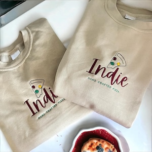 May include: Two beige sweatshirts with the text "Indie Hand Crafted Pizza" embroidered on them. One sweatshirt has a red and green pizza slice graphic, the other has a green and yellow pizza slice graphic.