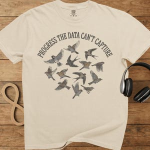 May include: Cream-colored t-shirt featuring a circular design of various birds in flight. The phrase "PROGRESS THE DATA CAN'T CAPTURE" is printed in a semi-circle above the birds. A pair of black headphones rests on the right.