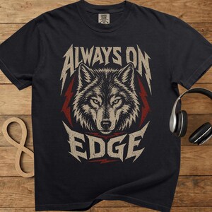 May include: Dark grey t-shirt with a wolf graphic and the words "ALWAYS ON EDGE". The design has a distressed, vintage look with red lightning bolts. Headphones are visible on the right side of the image.